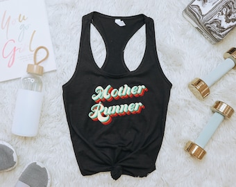 May include: Black tank top with the text "Mother Runner" in a retro font with green, red, and yellow colors. The tank top is on a white fuzzy surface.