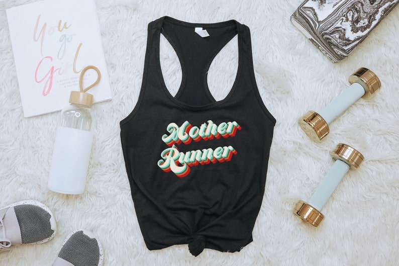 K&ouml;nnte beinhalten: Schwarzes Tanktop mit dem Text "Mother Runner" in einer Retro-Schriftart mit gr&uuml;nen, roten und gelben Farben. Das Tanktop liegt auf einer wei&szlig;en, flauschigen Oberfl&auml;che.