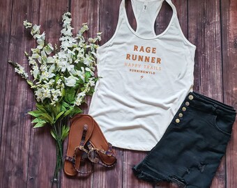 Könnte beinhalten: Eine Draufsicht mit einem weißen Racerback-Tanktop mit den Worten "Rage Runner Happy Trails Runningwyld" in Orange, kombiniert mit schwarzen Jeansshorts und braunen Sandalen. Weiße Blumen und grüne Blätter sind auf einer Holzoberfläche angeordnet.