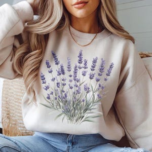 Op de afbeelding: Een cr&egrave;mekleurige trui met een paarse lavendelbloemprint. Het model draagt ​​een blauwe jeans en witte sneakers.