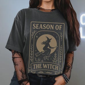 May include: Dark gray t-shirt featuring a beige graphic with the words "SEASON OF THE WITCH" and an illustration of a witch on a broomstick in front of a full moon. The design is framed by decorative elements.