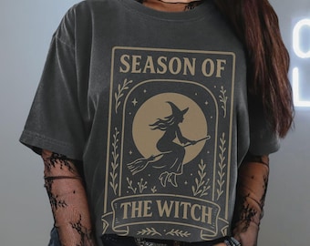 Puede incluir: Camiseta gris oscuro con un diseño beige. El diseño presenta el texto "SEASON OF THE WITCH" y una ilustración de una bruja en una escoba frente a una luna llena, enmarcada por elementos decorativos.