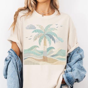 May include: Cream-colored t-shirt featuring a beach scene with a palm tree, waves, and clouds. The design uses patchwork-style patterns in shades of blue, green, and beige. The shirt has rolled-up sleeves and a relaxed fit.