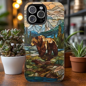 May include: A phone case with a stained glass design featuring a brown bear in a forest setting. The case has a blue, green, and brown color scheme.