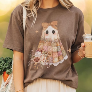 May include: A brown t-shirt featuring a whimsical ghost design. The ghost has a bow, floral patchwork dress, and a bouquet of flowers. The shirt is a neutral color and has a relaxed fit. The overall theme is a blend of spooky and floral.