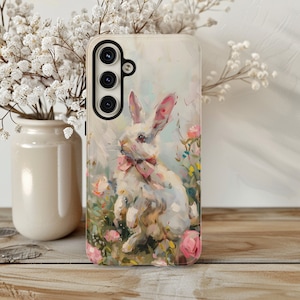 K&ouml;nnte beinhalten: Eine wei&szlig;e Handyh&uuml;lle mit einem gemalten Bild eines wei&szlig;en Kaninchens mit einer rosa Schleife in einem Feld mit rosa Blumen.