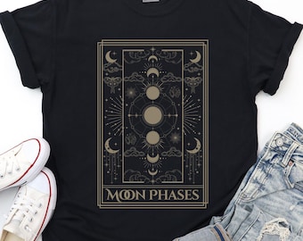 May include: Black t-shirt featuring a gold-colored graphic of moon phases, stars, and clouds. The text "MOON PHASES" is displayed in a rectangular frame. White sneakers and blue jeans are visible.