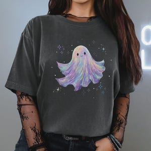 May include: Dark gray t-shirt featuring a colorful ghost illustration. The ghost is depicted with a smiling face and a flowing, pastel-colored body. The design includes stars and sparkles, creating a whimsical and playful aesthetic.