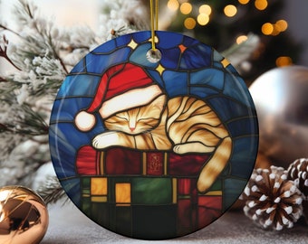 Puede incluir: Adorno navideño redondo con un gato atigrado durmiendo con un gorro de Papá Noel. El gato descansa sobre una pila de libros, sobre un fondo azul estilo vidriera con estrellas. Una cuerda dorada está unida para colgarlo.