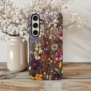 May include: A smartphone case featuring a colorful floral stained-glass design. The case showcases a variety of flowers in shades of purple, yellow, red, and orange, set against a brown background. The case is on a wooden surface.