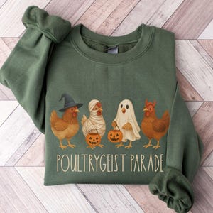 May include: Green sweatshirt featuring a Halloween-themed graphic. The design includes four chickens dressed as a witch, mummy, ghost, and devil, each holding a jack-o'-lantern. The text "Poultrygeist Parade" is printed below the chickens.