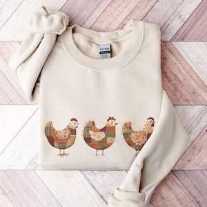 May include: A beige sweatshirt featuring three patchwork-style chicken designs in shades of brown, orange, and cream. The chickens have red combs and are arranged horizontally across the front of the garment. The sweatshirt has a crew neck and long sleeves.