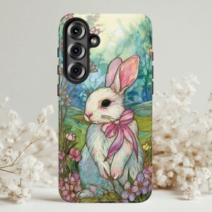 May include: A phone case featuring a watercolor illustration of a white rabbit with pink-lined ears and a pink bow. The rabbit is surrounded by pink and purple flowers, with a soft green and blue background.