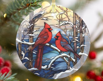May include: Round glass Christmas ornament featuring two red cardinal birds perched on a snow-covered branch. The ornament has a stained-glass style design with a winter landscape background, and a gold ribbon for hanging.