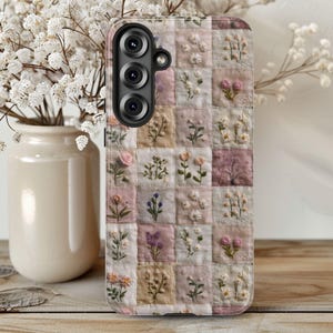 May include: A phone case featuring a patchwork design with embroidered floral motifs in pink, cream, and beige squares. The case has a black camera module with three lenses and is set against a blurred background with a vase of white flowers.