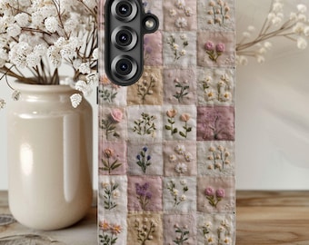 以下が含まれることがあります： ピンク、クリーム、ベージュの正方形に刺繍された花のモチーフがパッチワーク模様になった携帯電話ケース。ケースには3つのレンズが付いた黒いカメラモジュールが付いています。背景はぼやけており、白い花の入った花瓶があります。