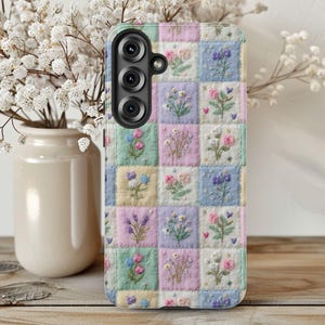 May include: A phone case with a patchwork design featuring embroidered flowers in pastel colors. The case has a black camera module and a soft, quilted texture. The floral squares include pink, blue, yellow, and green hues.