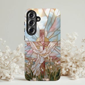 May include: A phone case featuring a stained-glass design with a cross, sun, and floral elements. The case has a black camera module and a colorful, artistic depiction of a religious scene with pastel hues.