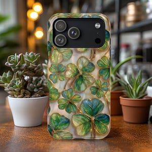 May include: A phone case featuring a repeating pattern of green and blue shamrocks with gold-colored outlines. The case has a glossy finish and a camera module with three lenses. The background is a cream color.