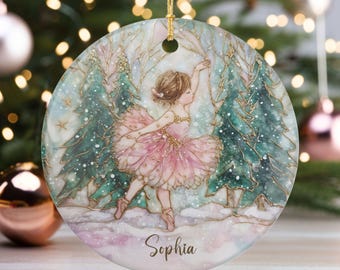 May include: A round Christmas ornament featuring a ballerina in a pink tutu dancing in a snowy forest scene. The ornament has a gold string for hanging and the name "Sophia" is written on the bottom. The background includes Christmas trees and a bokeh effect.