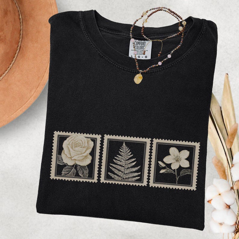 May include: Black t-shirt featuring three postage stamp designs with botanical illustrations: a rose, a fern, and a flower. A beaded necklace with a pendant rests on the shirt. The shirt is made of a soft, comfortable material.