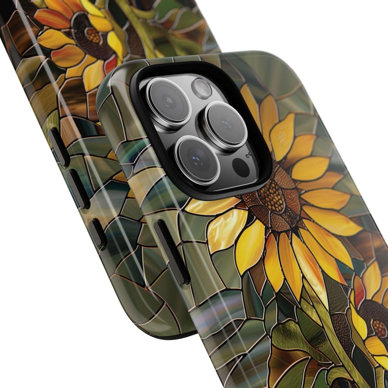 May include: A phone case with a stained glass design featuring sunflowers in shades of yellow, brown, and green. The case has a black camera cutout and a raised lip around the edges.