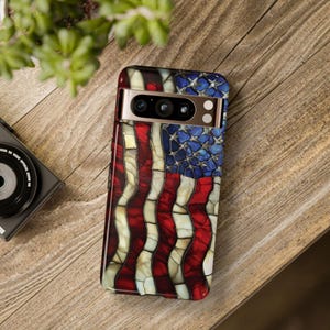 May include: A smartphone case featuring a stained-glass design of the American flag. The case has a red, white, and blue color scheme, with a gold-colored camera module. The case is on a wooden surface.