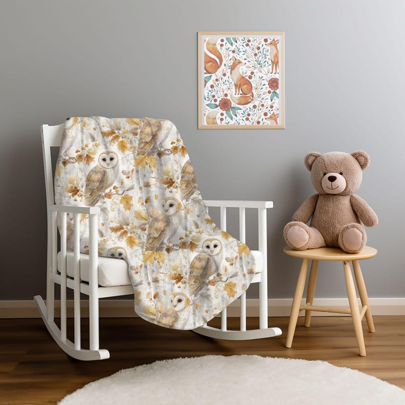 May include: A white rocking chair draped with a blanket patterned with owls and autumnal leaves. A framed print with foxes and floral designs is on the wall. A brown teddy bear sits on a small wooden stool, creating a nursery scene.