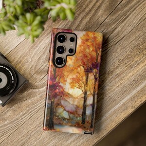 May include: A smartphone case featuring a landscape painting of trees with orange and red leaves, set against a sunset. The case has a glossy finish and a multi-lens camera cutout. The case is on a wooden surface.