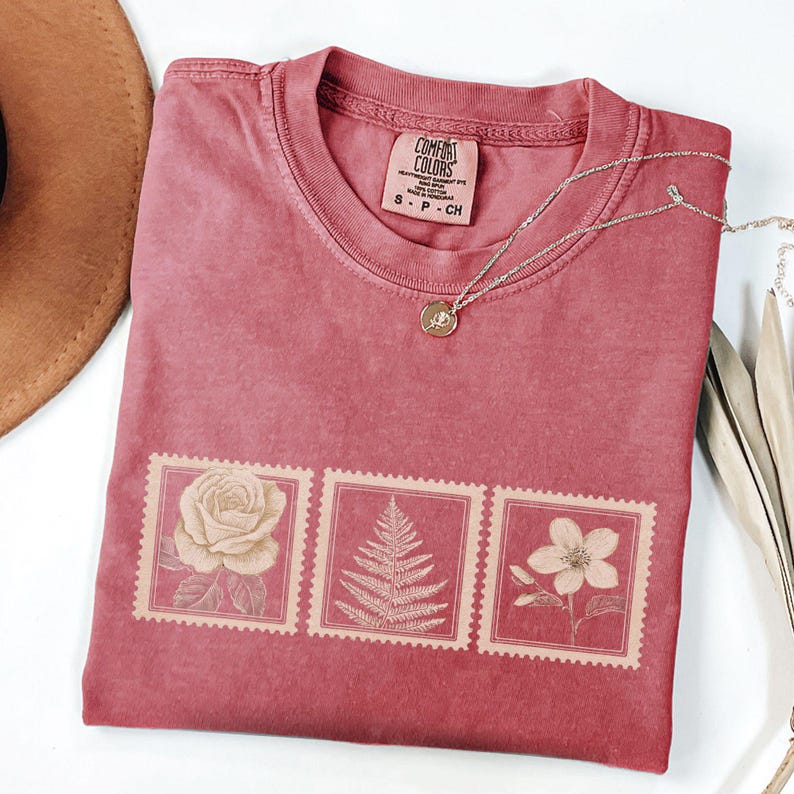 May include: A dusty rose Comfort Colors t-shirt featuring three postage stamp designs with botanical illustrations. The stamps depict a rose, fern, and flower. A silver necklace with a small pendant rests on the shirt.