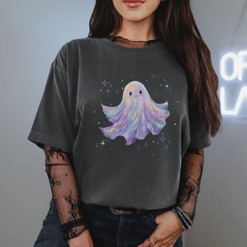 May include: Dark gray t-shirt featuring a colorful ghost illustration. The ghost is depicted with a smiling face and a flowing, pastel-colored body. The design includes stars and sparkles, creating a whimsical and playful aesthetic.