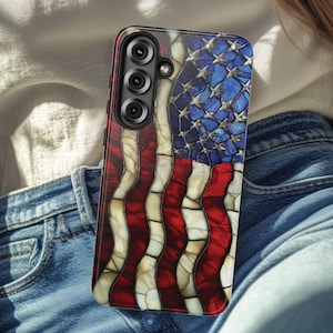 May include: A smartphone case featuring a stained-glass design of the American flag. The case has a red, white, and blue color scheme, with a detailed pattern of stars and stripes. The case has a black camera module with three lenses.