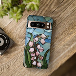 Pu&ograve; includere: Una custodia per telefono con un design in vetro colorato che presenta fiori di mughetto rosa su uno sfondo blu e verde.