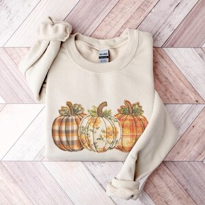 May include: A cream-colored sweatshirt featuring three decorative pumpkins. The pumpkins have different patterns, including plaid and floral designs, in shades of orange, brown, and yellow. The sweatshirt has a crew neck and long sleeves.