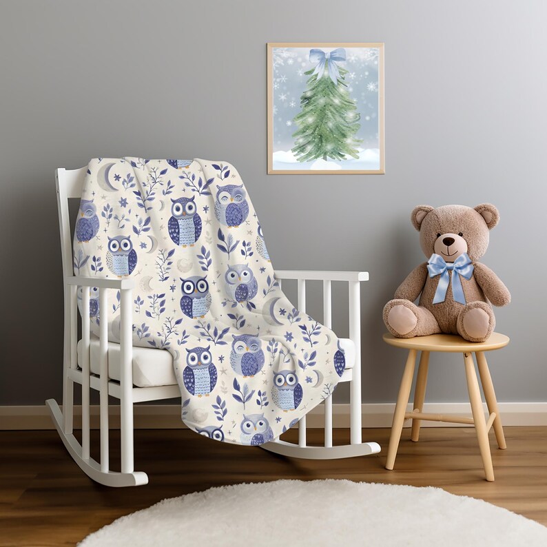 May include: A cream-colored throw blanket with a blue owl pattern. The blanket is draped over a white rocking chair with a teddy bear sitting on a wooden stool in the background.