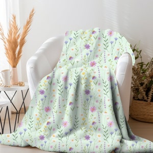 May include: A soft, green throw blanket with a floral pattern featuring pink, purple, and yellow flowers. The blanket is draped over a white armchair.