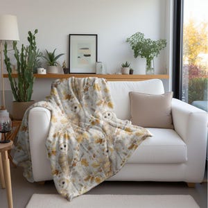 May include: A white sofa with a beige cushion and a decorative throw. The throw features a pattern of owls, leaves, and branches in brown, beige, and gold. The blanket is draped over the sofa, creating a cosy atmosphere.