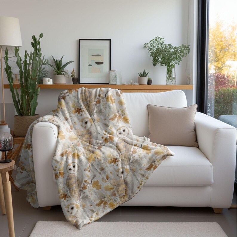 May include: A white sofa with a beige cushion and a decorative throw. The throw features a pattern of owls, leaves, and branches in brown, beige, and gold. The blanket is draped over the sofa, creating a cosy atmosphere.