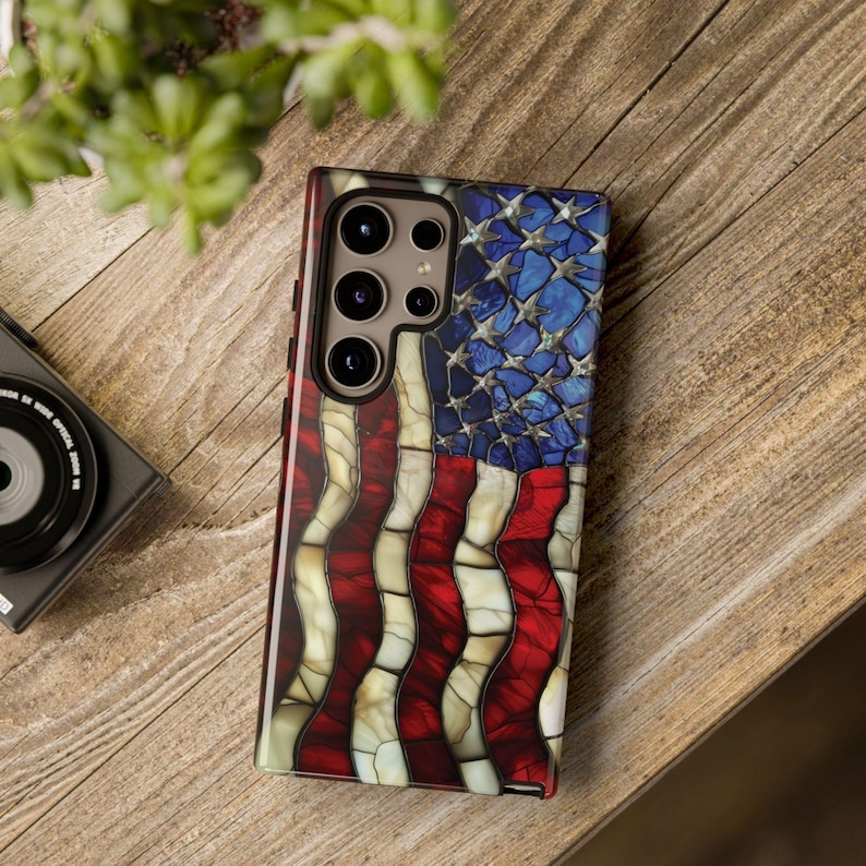 May include: A smartphone case featuring a mosaic-style American flag design. The case has a glossy finish with red and white stripes and a blue field of stars. The case is shown on a wooden surface.