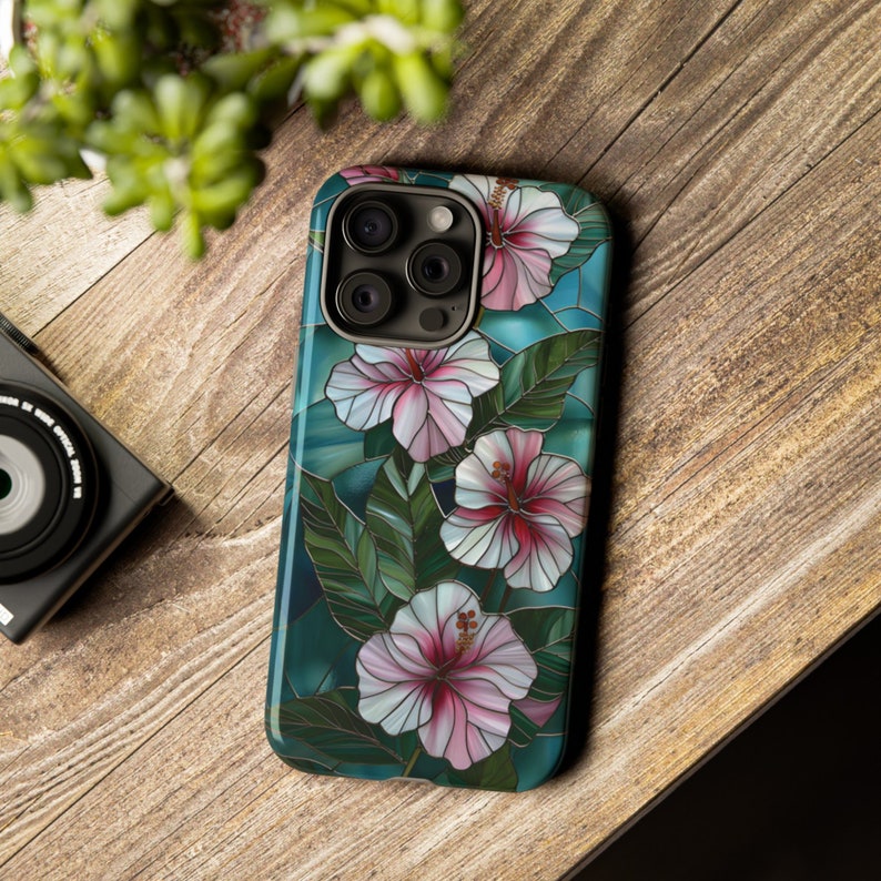 May include: A phone case with a stained glass design featuring pink and white hibiscus flowers on a teal background.