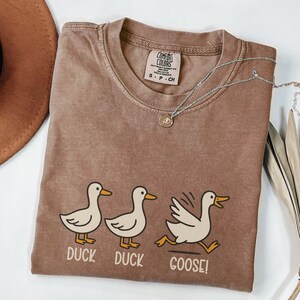 May include: Brown t-shirt with a crew neck and the words "DUCK DUCK GOOSE!" below a cartoon duck graphic. The shirt has a Comfort Colors tag and is paired with a silver necklace. The shirt is size S-P-CH.