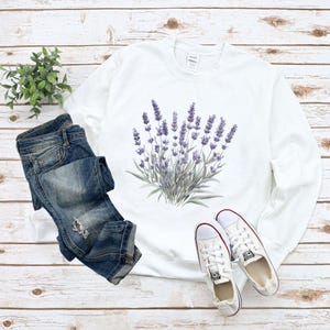 Op de afbeelding: Witte trui met een lavendelbloemenprint. De trui is gecombineerd met een blauwe jeans en witte sneakers.
