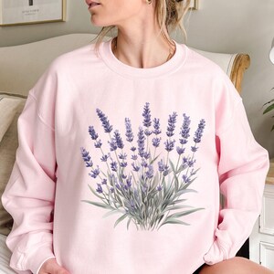 Op de afbeelding: Een roze sweatshirt met een lavendelbloemendruk. De bloemen zijn paars en groen.
