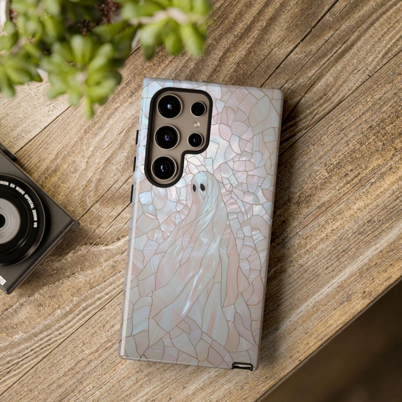 May include: A white phone case with a pearly iridescent design and a ghost figure. The case has a camera lens cutout with three lenses.