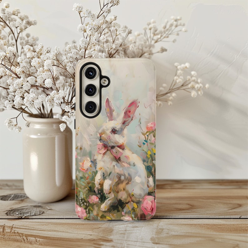 K&ouml;nnte beinhalten: Eine wei&szlig;e Handyh&uuml;lle mit einem gemalten Bild eines wei&szlig;en Kaninchens mit einer rosa Schleife in einem Feld mit rosa Blumen.