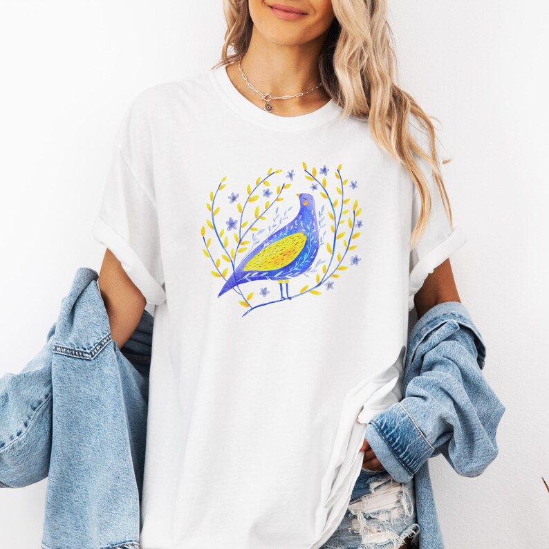 Puede incluir: Camiseta blanca con una ilustraci&oacute;n de un p&aacute;jaro azul y amarillo rodeado por una corona de hojas amarillas y flores azules. El p&aacute;jaro tiene el pecho amarillo y las alas azules. La camiseta tiene mangas enrolladas.