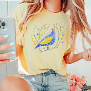 Puede incluir: Camiseta amarilla clara con un dise&ntilde;o de p&aacute;jaro azul y amarillo rodeado de elementos florales amarillos y azules. El p&aacute;jaro tiene un aspecto estilizado. La camiseta est&aacute; hecha de un material suave y es adecuada para ropa casual.