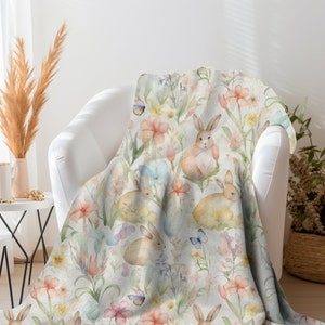 May include: A soft, white throw blanket with a watercolor pattern of bunnies and flowers. The bunnies are brown and white, and the flowers are pink, yellow, and blue.