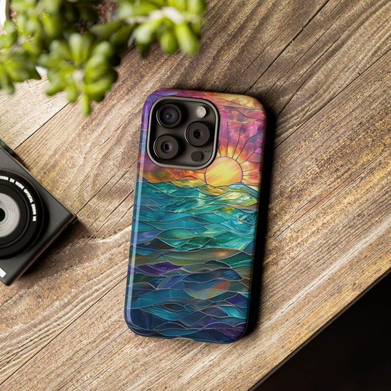 Pu&ograve; includere: Una custodia per telefono colorata con un designo a vetri colorati di un tramonto sull'oceano. La custodia ha un ritaglio per la fotocamera con tre obiettivi.