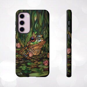 May include: A phone case with a stained glass design featuring a green frog with red eyes and pink flowers. The case is black and has a cutout for the camera lens.